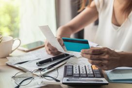 女人在办公桌前用计算器用信用卡支付她每月的账单＂width=