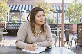 一名女子坐在户外咖啡馆里，在笔记本上写字