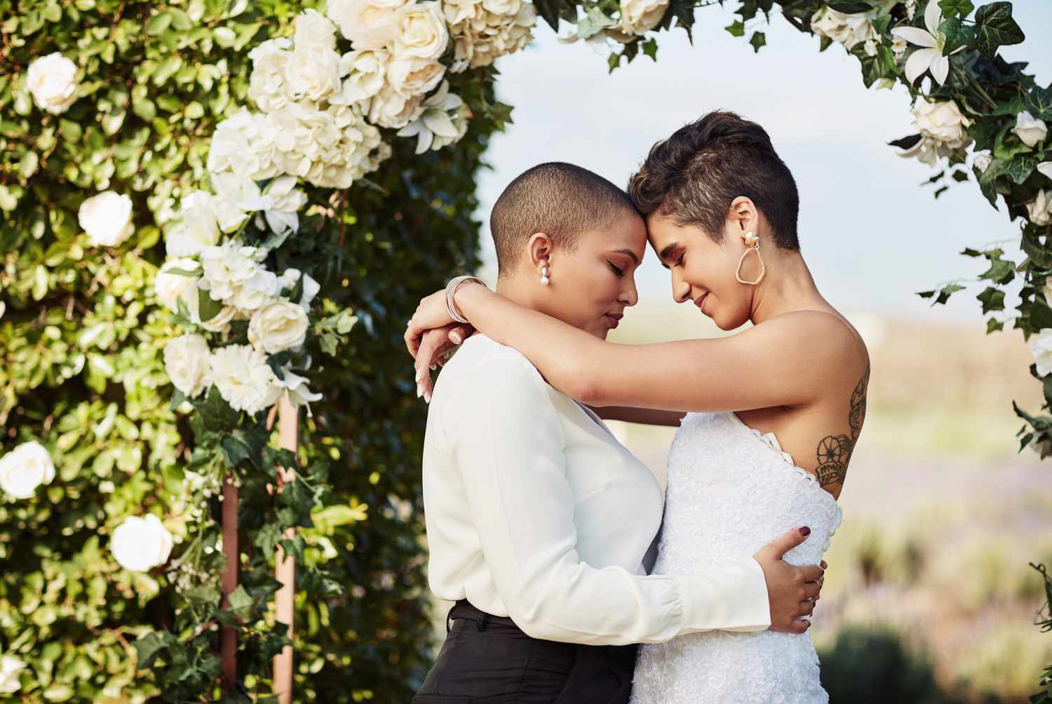 一对女同性恋夫妇拥抱在自己婚礼的衣服站在花前拱”class=