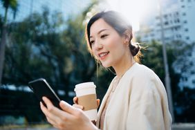 自信的年轻亚洲女商人一边阅读电子邮件，一边喝着咖啡，她走在城市的金融区，在清新的早晨，对面是商业建筑