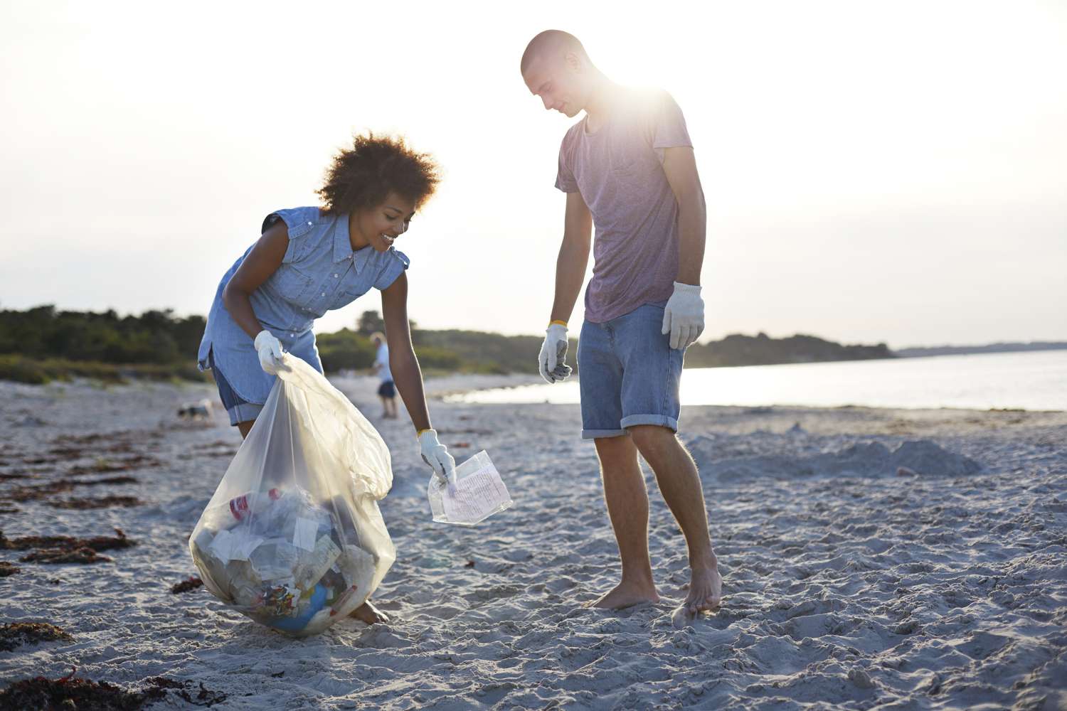 两个人站在海滩上捡垃圾，把它放进塑料袋＂class=