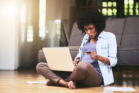 一名黑人妇女坐在沙发前的硬木地板上，手里拿着笔记本电脑和信用卡。＂>
          </noscript>
         </div>
        </div>
       </div>
       <div class=
