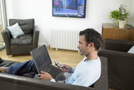 在一间开着电视的客厅里，一名男子坐在那里，膝上放着一台笔记本电脑，手里拿着电视遥控器