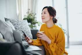 Woman working on smartphone and laptop with coffee mug in hand