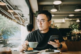 一个年轻人坐在咖啡桌旁，凝视着他右边的窗户。他右手拿着一杯茶，左手拿着一部智能手机。