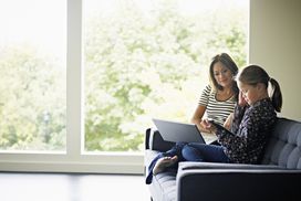 在现代家庭中，母亲坐在沙发上，膝上放着笔记本电脑，看着女儿在平板电脑上工作＂width=